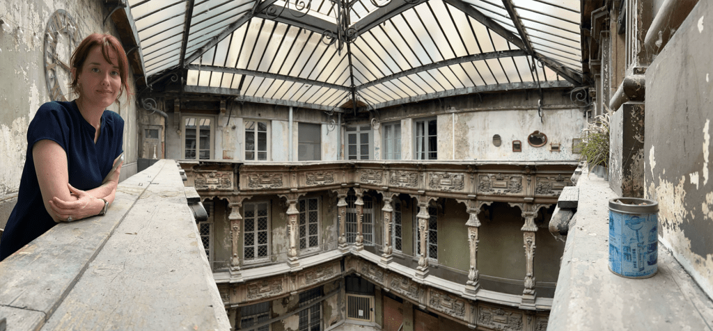 A woman stands on a balcony in a partially derelict building, showcasing intricate architectural details and a glass roof.