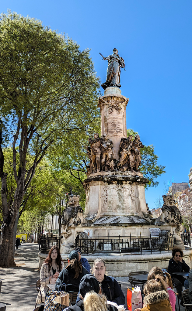 A statue atop a monument stands in a park with people seated at tables below it, surrounded by trees and a blue sky.