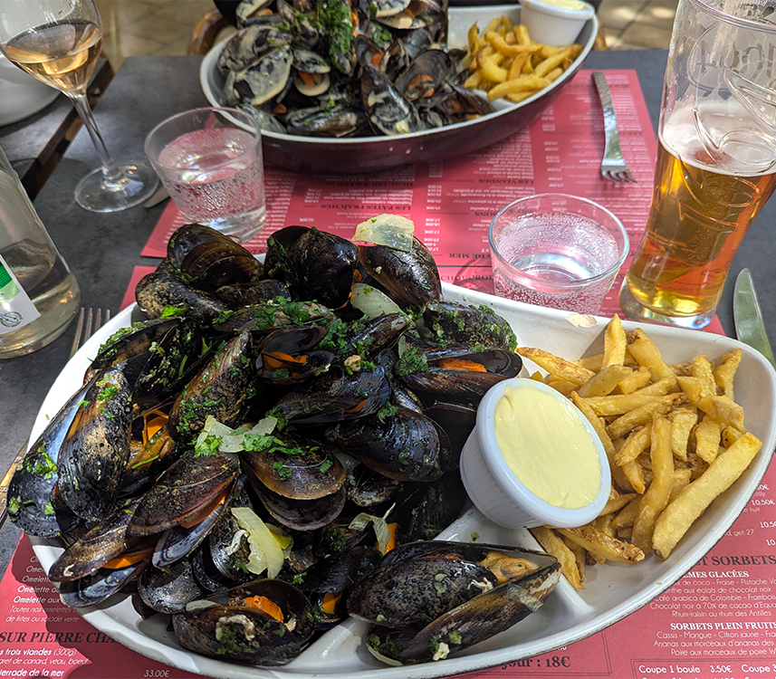 A plate of mussels served with fries and a small dish of sauce, with drinks and a menu in the background.