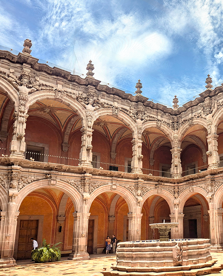 Baroque architecture of the former convent of San Agustin, featuring arched colonnades, intricate stone carvings, and a central fountain, under a blue sky.
