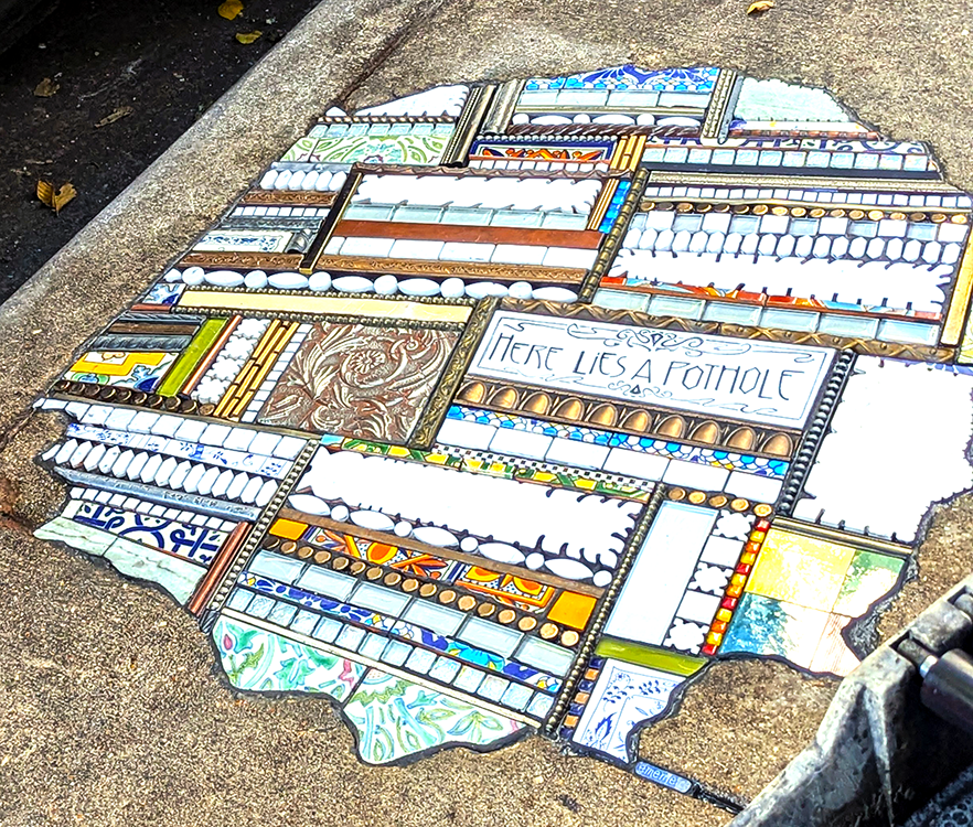 Colorful mosaic artwork reading 'Here Lies a Pothole,' installed in a sidewalk as a creative repair by the street artist Ememem.