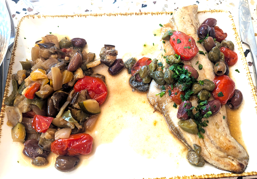A plate featuring grilled fish garnished with olives, capers, and cherry tomatoes, accompanied by a medley of sautéed vegetables including eggplant, bell peppers, and zucchini.