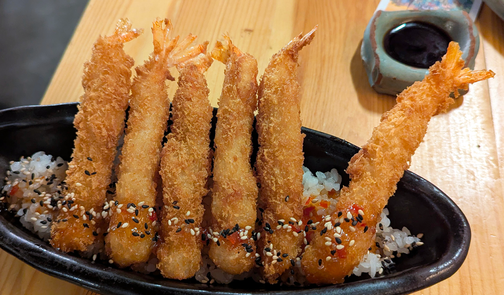 A serving of crispy golden shrimp tempura on a bed of sushi rice, garnished with black and white sesame seeds.