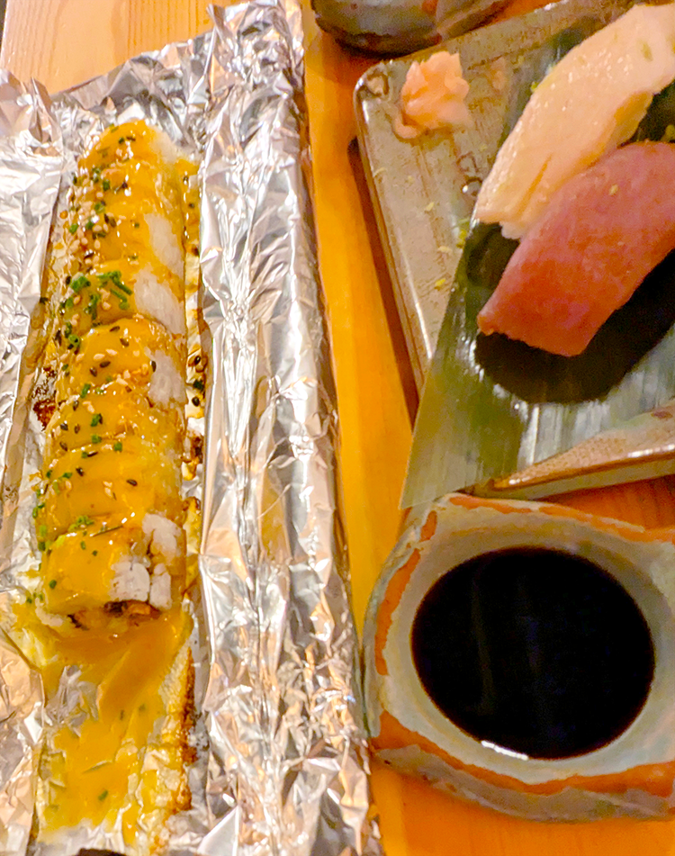 A plate of sushi featuring a rolled sushi with a yellow sauce, garnished with sesame seeds and chives, next to a selection of sashimi including tuna and white fish, with pickled ginger and soy sauce on the side.
