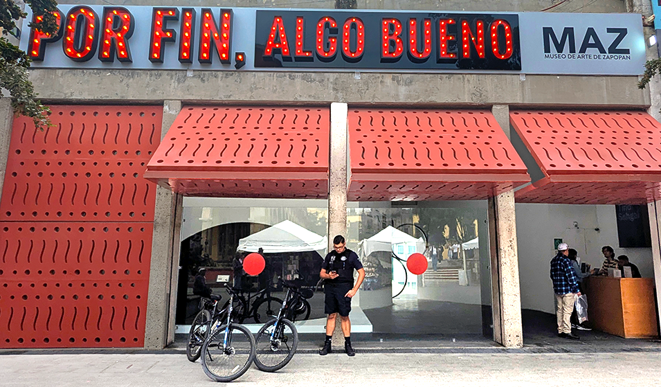 Exterior view of the Museo de Arte de Zapopan (MAZ) featuring a bright sign that reads 'POR FIN, ALGO BUENO', with a security guard and bicycles in front.