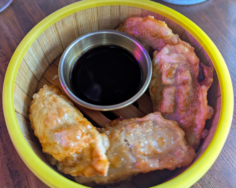 A bamboo steamer with four golden-brown dumplings and a small bowl of dark dipping sauce.