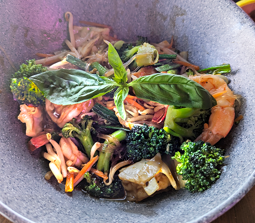 A close-up of a bowl of stir-fried noodles with shrimp, broccoli, green peppers, and bean sprouts, garnished with fresh basil leaves.