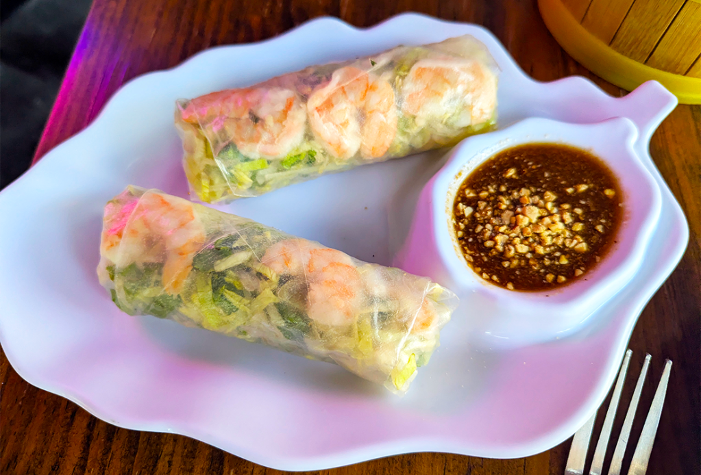 Two shrimp spring rolls on a white plate, accompanied by a small dish of dipping sauce topped with crushed peanuts.