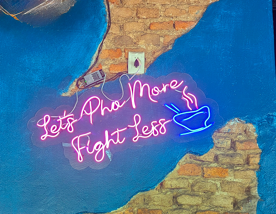 Neon sign reading 'Let's Pho More Fight Less' against a colorful textured wall.