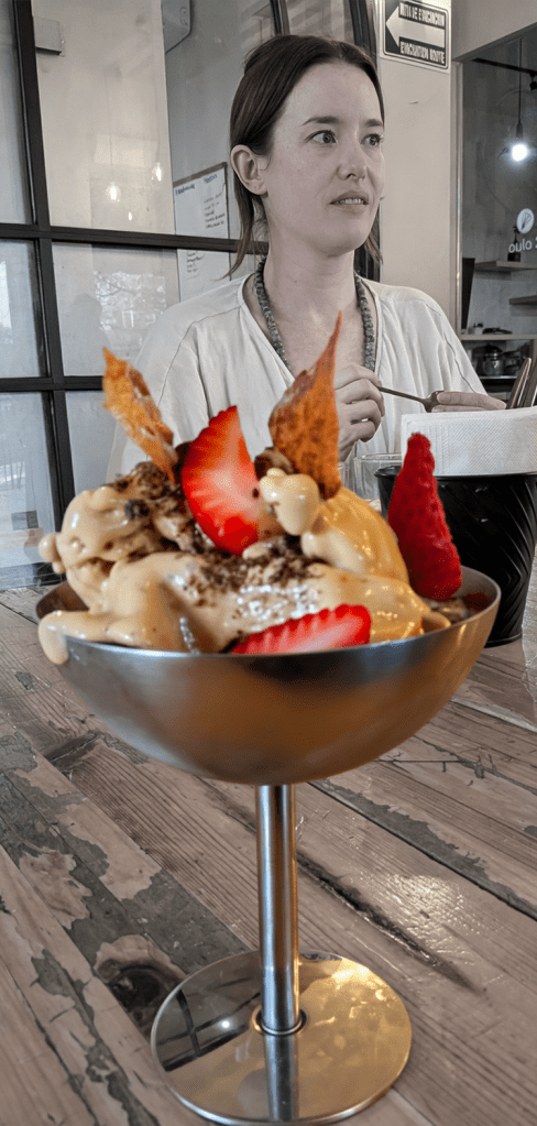 A close-up of a dessert served in a metallic bowl, topped with strawberries and crispy decorations, with a woman seated in the background.